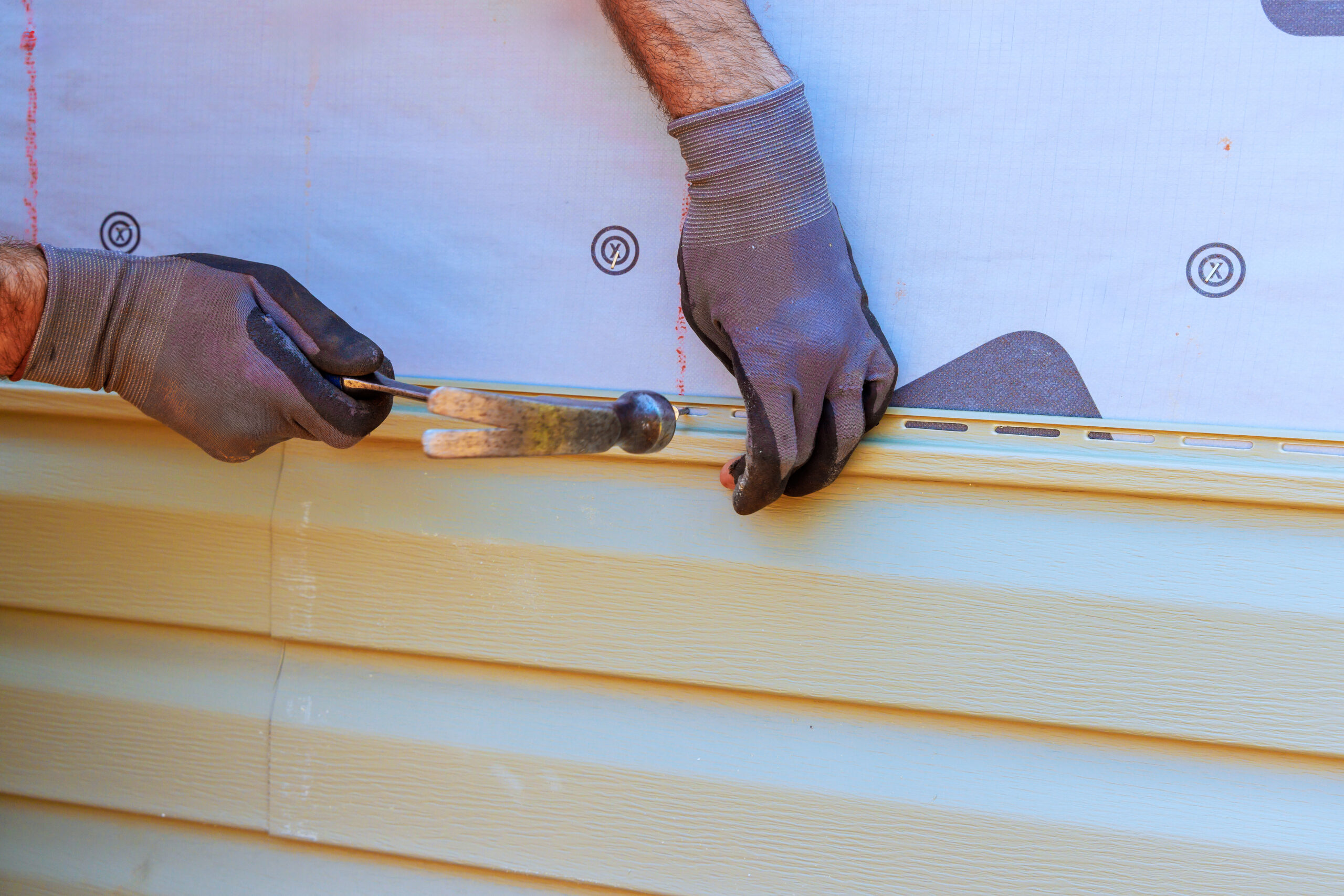 Worker carefully installs vinyl siding on house, ensuring level alignment while using hammer tools.