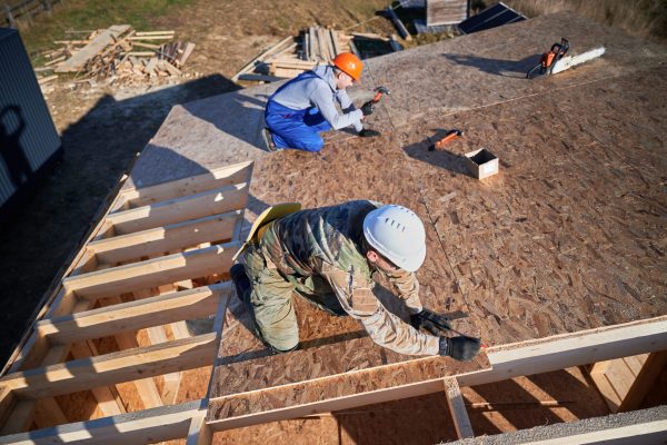 Carpenters hammering nail into OSB panel on the roof top of future cottage. Men workers building wooden frame house. Carpentry and construction concept.