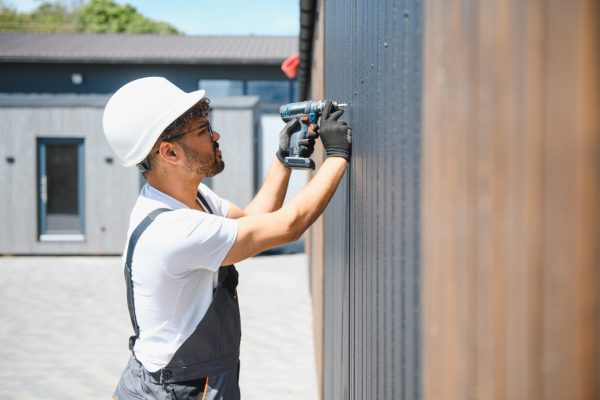 Construction worker using a cordless drill installing siding on a prefabricated building on a sunny day